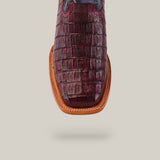 Close-up of a brown, square-toed shoe with a textured crocodile pattern similar to Exotic Caiman Belly Tail boots, featuring a tan trim. The sole subtly casts a shadow on the beige background.