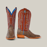 The Exotic Caiman Belly Tail cowboy boots feature gray, textured leather bottoms and reddish-brown shafts with intricate blue and white stitching. They include wooden heels and a subtle blue lining for added flair.