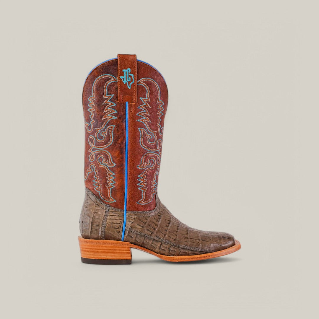 Discover our Exotic Caiman Belly Tail cowboy boot in a sleek gray square toe design. Featuring a beautiful alligator-skin-like leather finish, it has intricate red and blue embroidery, a striking blue logo pull tab, and a sturdy wooden heel on a sophisticated beige background.