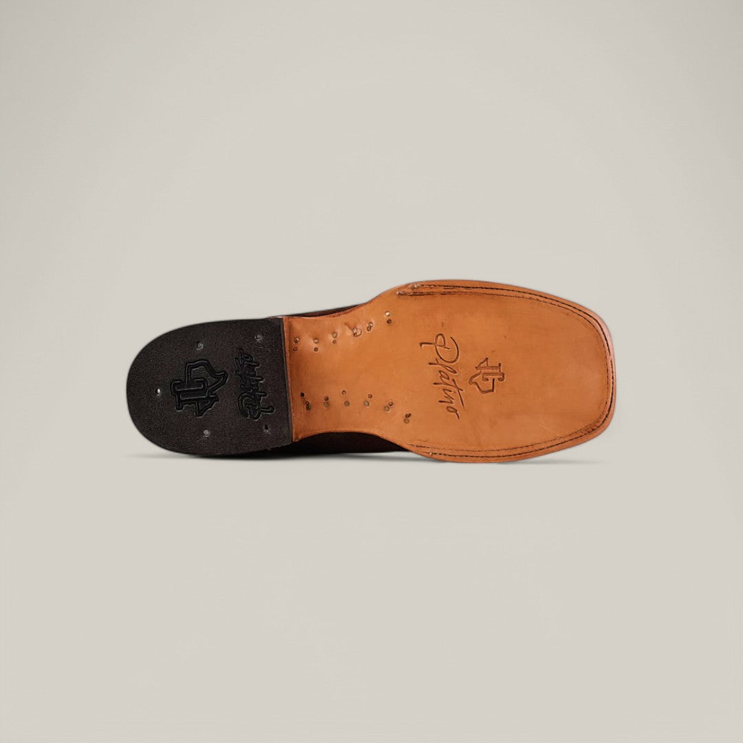 The image showcases the sole of the Brown Suede - Platinum - Square Toe cowboy boot, placed horizontally. Made from tan leather with a square toe and darker heel, it features decorative stitching and embossed logos. A plain, light background enhances its rustic charm.