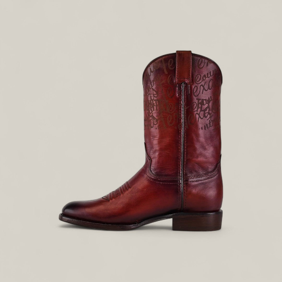 The Wyatt - Cogñac - Roper Toe is a single, mid-calf brown cowhide boot with decorative stitching and engraved text on the shaft, shown in profile against a plain light background to highlight its quality craftsmanship.
