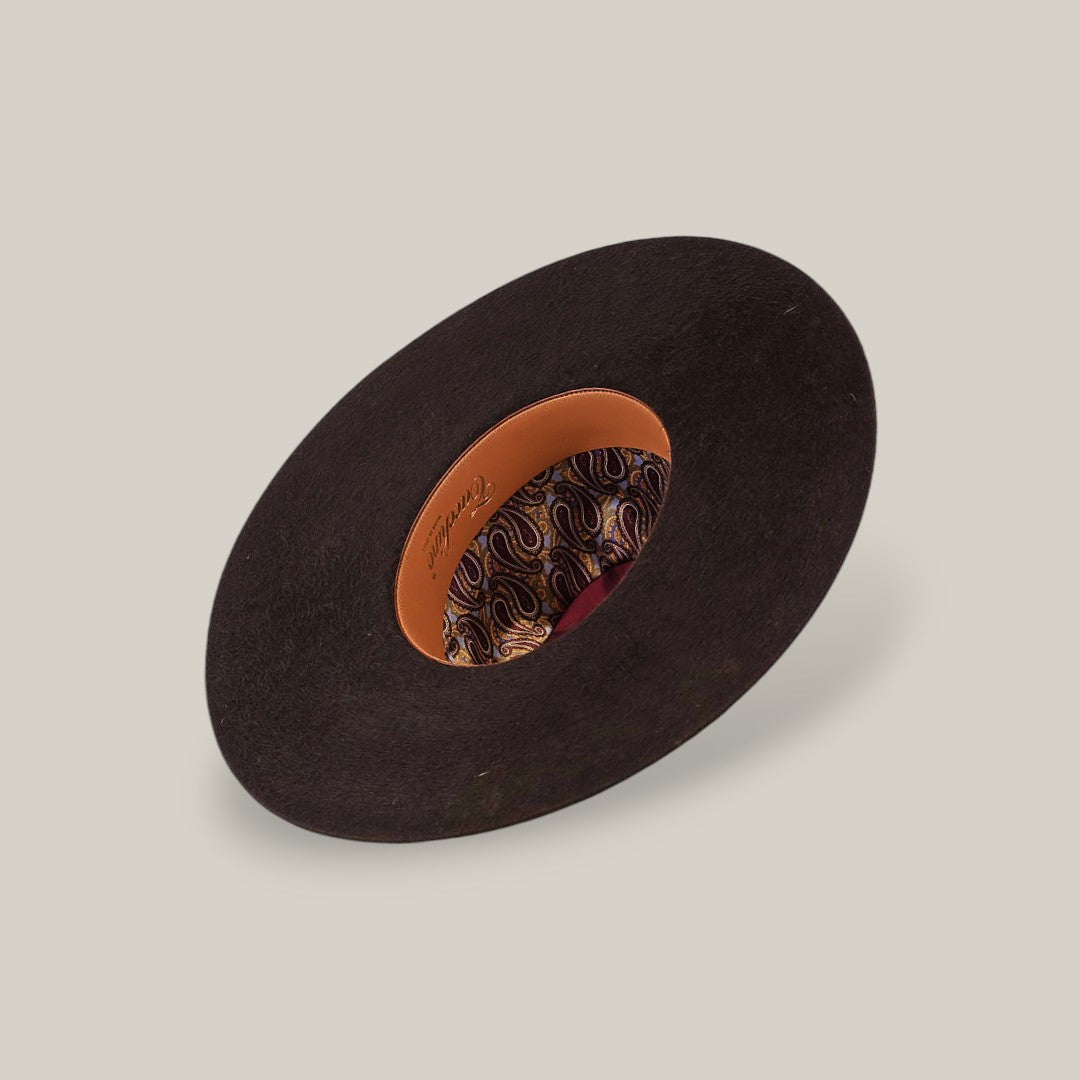The 10X Grizzly - Tacchino luxury hat, with a wide black brim, is displayed upside down to reveal its brown leather inner band and paisley-patterned lining against a plain beige background.