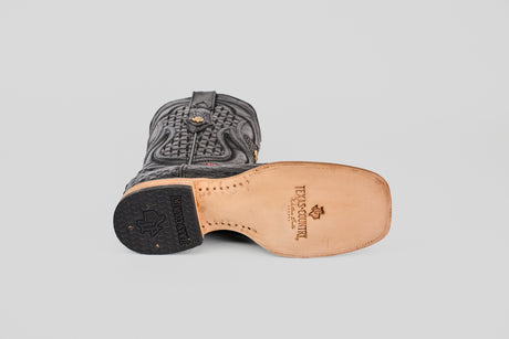 A single Exotic Caiman Horn Back - Black - Square Toe boot lies on its side, displaying the tan leather sole stamped with TEXAS COUNTRY and a decorative logo. The boot features a black heel and intricate stitching.