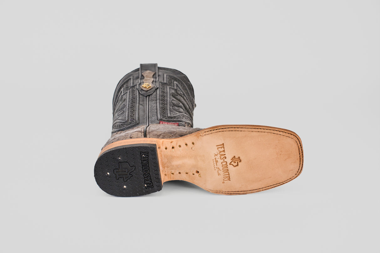 The Exotic Caiman Horn Back - Serpentine - Square Toe boot lies on its side, displaying detailed shaft stitching, a tan leather sole with the Texas Grip™ logo, a black heel, and a new-look square toe design.