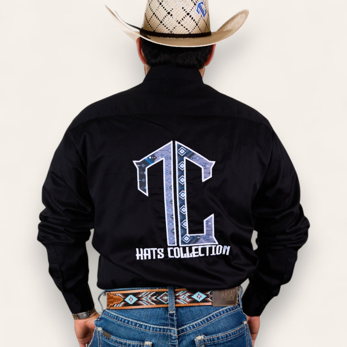 A man in a cowboy hat, black shirt with a large TC Hats Collection logo on the back, and blue jeans wears the TC Hats - Black Blue Aztec belt, featuring a stylish white and blue Aztec design, standing with his back to the camera.