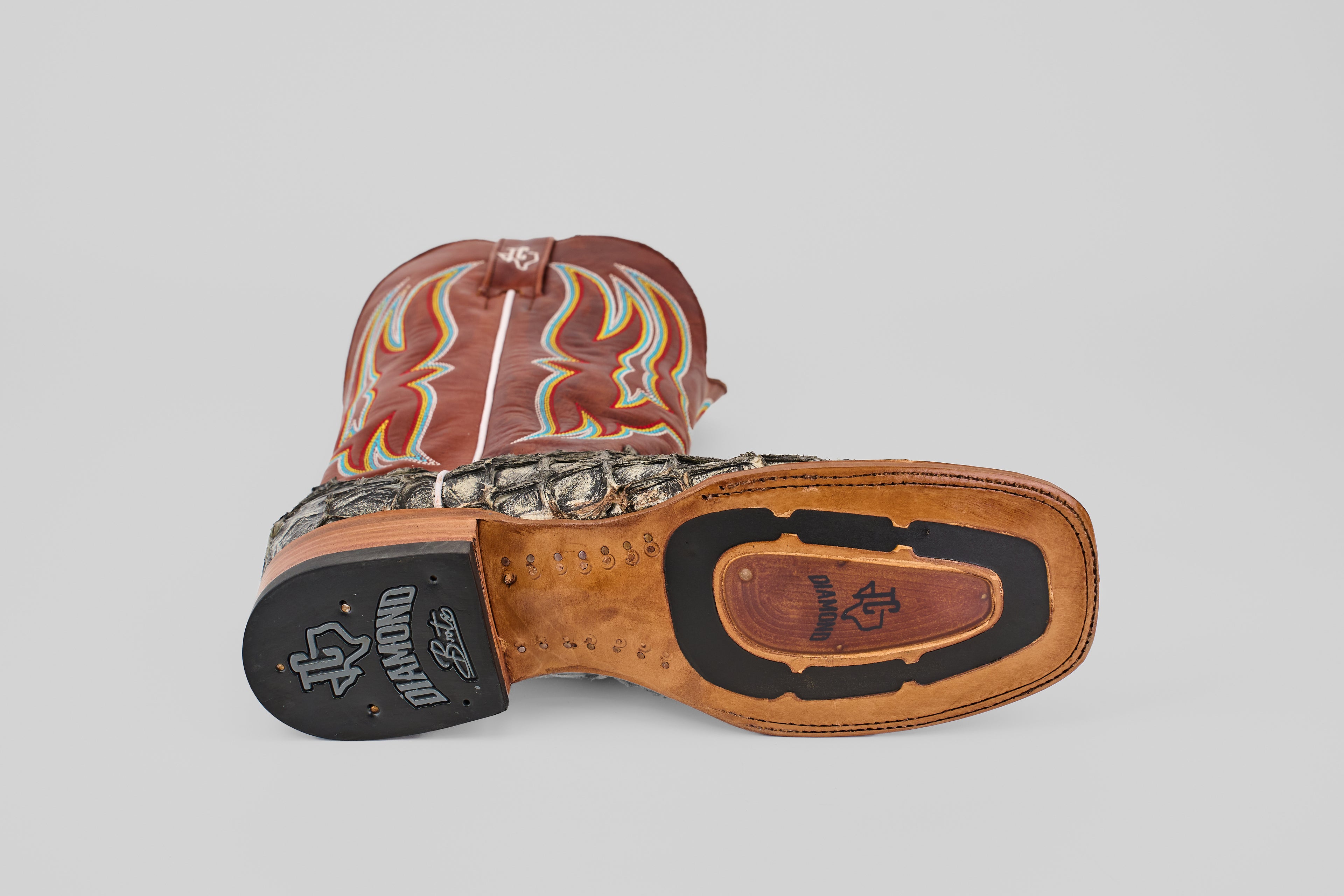 The Robinson - Newspaper - Square Toe boot is shown on its side, revealing a leather sole with black rubber grip, a handcrafted scale-textured Purarucu leather lower, and a brown upper accented by red and yellow stitching.