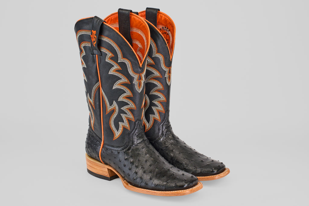 The Steele - Black - Square Toe boots, crafted from luxury ostrich leather with black and orange accents, showcase a textured pattern and wooden soles, positioned upright on a light gray background.