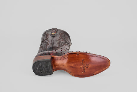 A handcrafted Exotic Full Quilt Ostrich - Black Cherry - Round Toe cowboy boot lies on its side, displaying detailed stitching, branded sole with metal studs along the arch. The background is plain light gray.