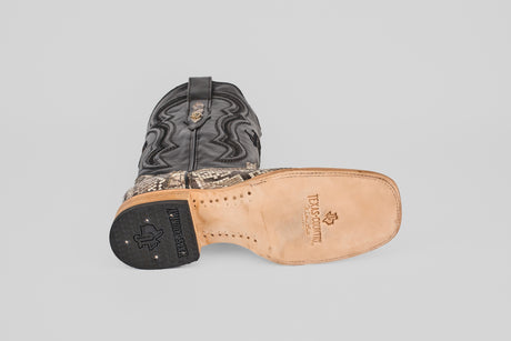 The Tanner - Natural - Square Toe cowboy boots feature black tops and Python Natural-patterned lowers. One boot stands upright while the other is on its side, displaying a tan leather sole with Texas Country branding and a black heel.