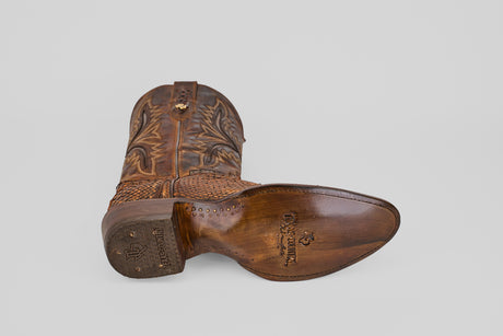 The Tanner – Chestnut – Round Toe boot rests on its side, displaying its detailed stitching and Lucchese-stamped sole. Set against a plain light gray background, it perfectly highlights classic western fashion style.