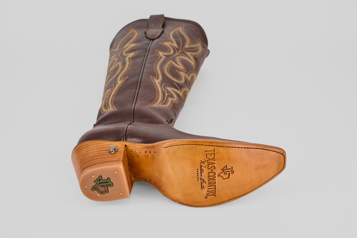 The Allison Dark Moka boot features dark brown leather, decorative stitching on a tall shaft, a tan sole with the Texas Country logo and “Handmade” stamp, plus a snip toe and angled heel for classic women’s cowboy style.