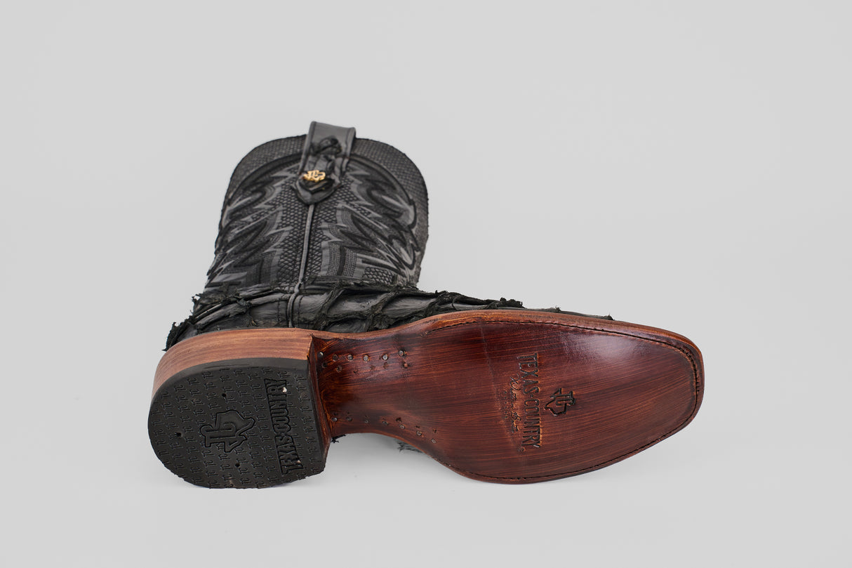 The Conor - Matte Black - Narrow Square Toe boot lies on its side, revealing “Handcrafted” engraved on the wooden heel and arch, with intricate stitching on the leather upper.