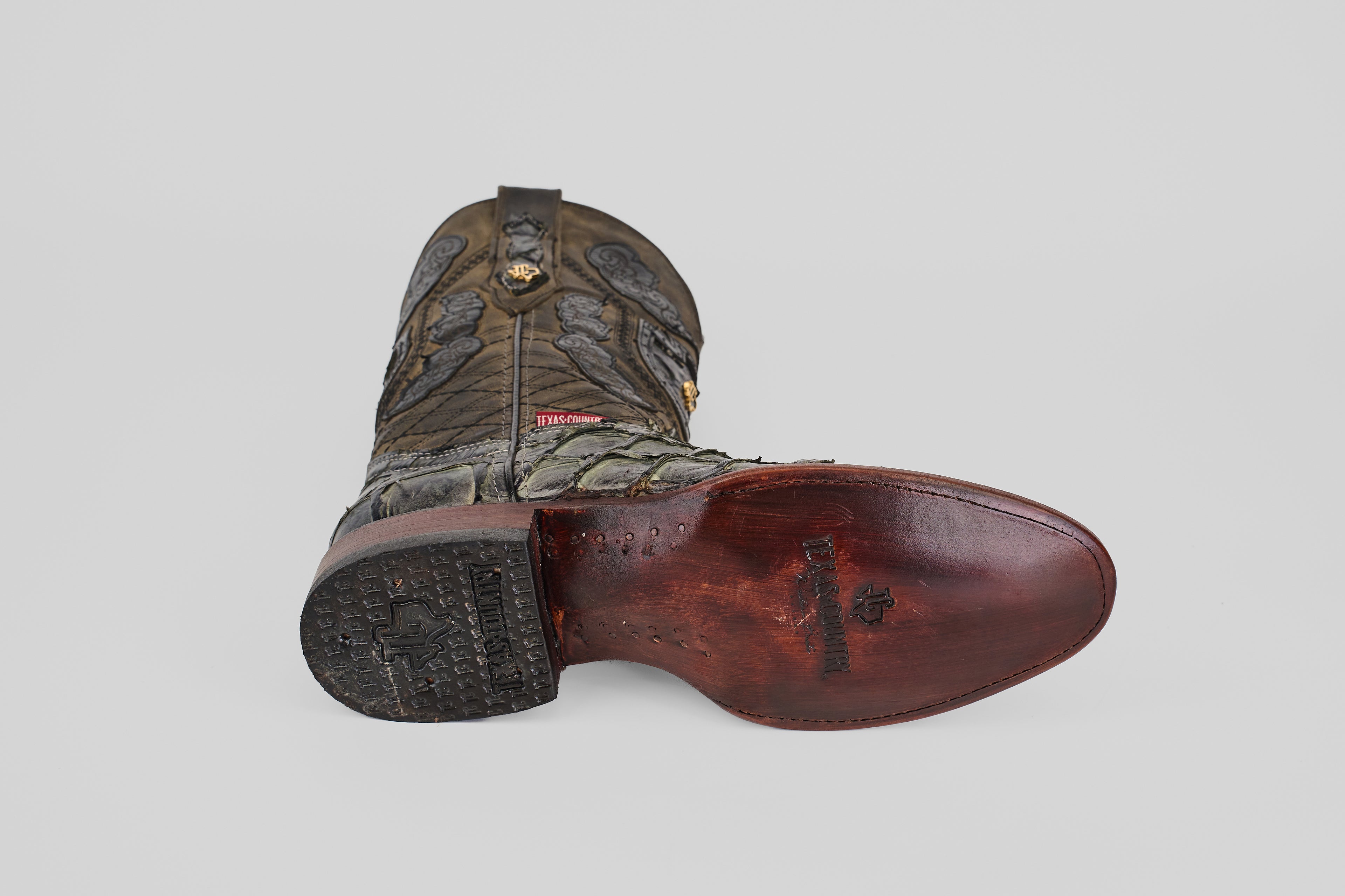 A single Conor - Charcoal - Roper Toe boot lies on its side, showcasing intricate upper stitching and a polished, reddish-brown fish leather sole with a decorative stamp and textured heel.