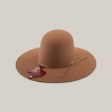 The 4X - Fawn - TC Hats is a light brown felt Western hat with a wide brim, round crown, and thin matching hatband. A maroon Africa-shaped tag hangs from the band. The hat is shown against a plain beige background.