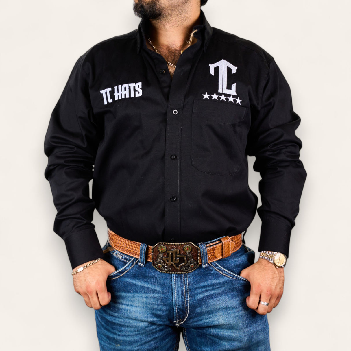 A man models the TC Hats - Black & White shirt with logo embroidery, paired with blue jeans, a brown tooled leather belt, gold watch, and jewelry against a plain white background.