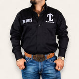 A man models the TC Hats - Black & White shirt with logo embroidery, paired with blue jeans, a brown tooled leather belt, gold watch, and jewelry against a plain white background.