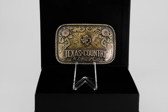 The Texas Country Buckle - Mod 1 is a decorative rectangular Western belt buckle with gold and silver floral designs, engraved with Texas Country Buckles, shown upright on a clear stand against a black background.