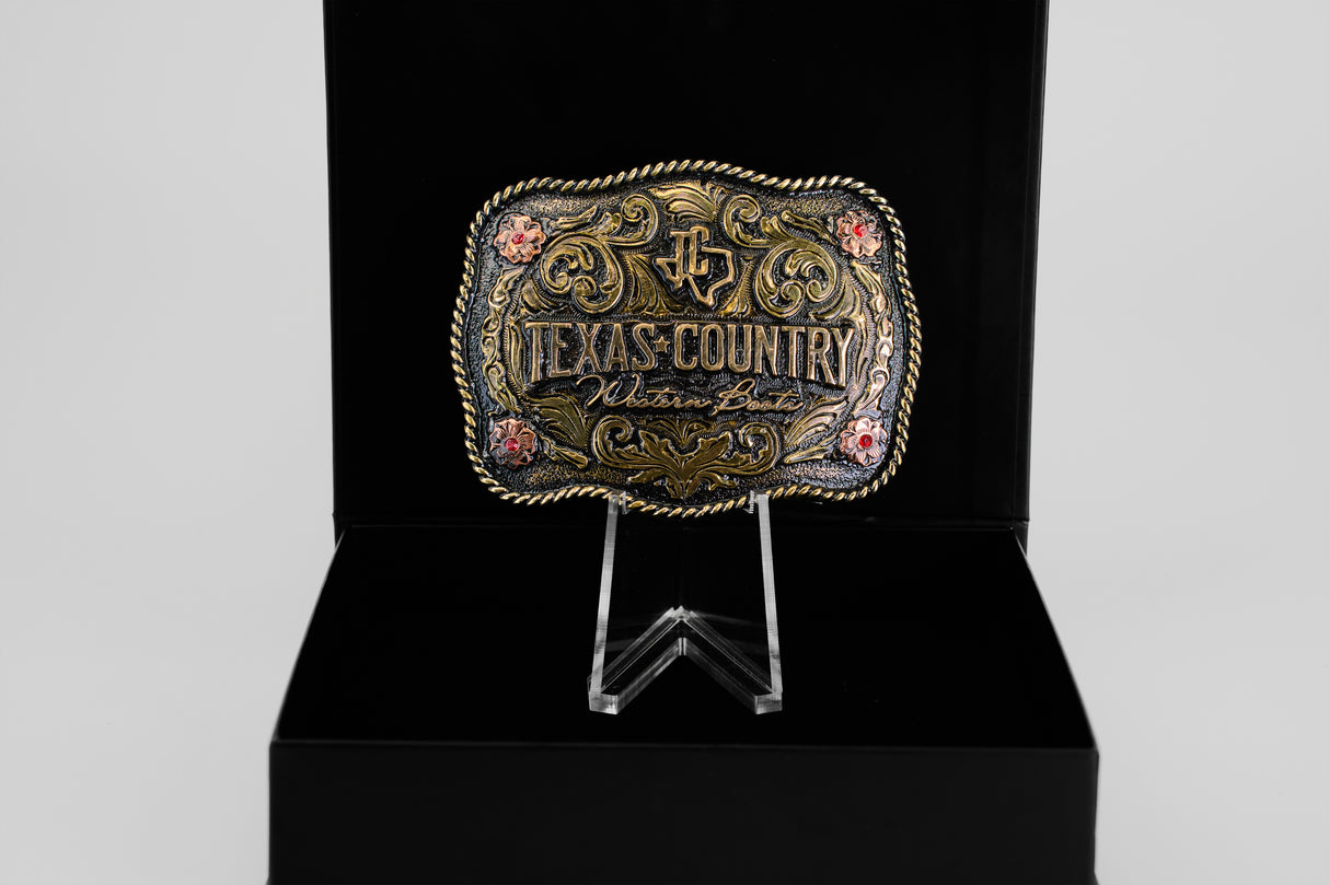 The Texas Country Buckle - Mod 5 is a gold and black belt buckle with floral accents, displayed on a stand in a black box. Engraved with Texas Country Western Best, its perfect for Western buckle collectors.