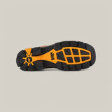 The image displays the durable sole of the Amazonas Brown Composite Toe - E6002 boot, featuring a black and orange tread pattern with a central logo and geometric shapes for enhanced grip. The backdrop is plain light gray.