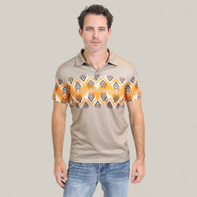 A man stands against a plain light gray background, wearing the Mens Performance Fabric Aztec Panoramic Print Khaki Polo (PGP10607) with a yellow and black geometric design across the chest and sleeves, paired with blue jeans.