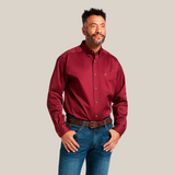 A man confidently stands with hands on hips in a Solid Twill Classic Fit Shirt Burgundy - 10012635, paired with blue jeans and a brown belt. The shirt features a chest pocket logo, while he smiles slightly against the plain white backdrop.