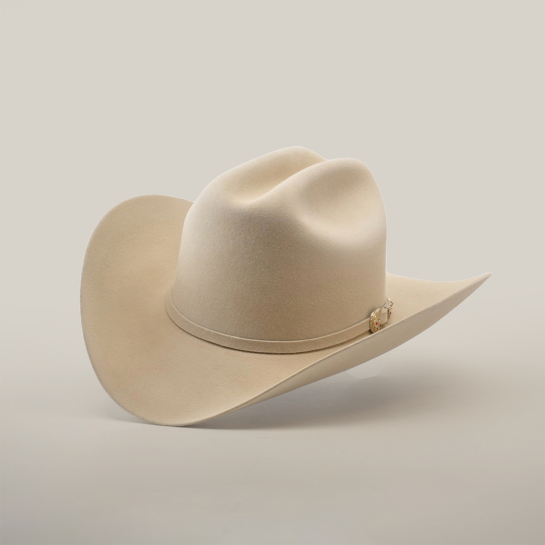The 6X Real Belly, a light beige fur felt hat with a wide brim and decorative band, rests on a smooth white surface. Its premium Western design features a high crown with two slight dents that enhance its classic style.