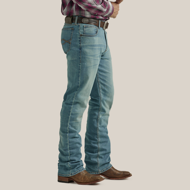 A person in Wrangler 20X Mens Light Wash Shade Bootcut Stretch Jeans - 112335407, brown leather cowboy boots, a brown belt, and a red plaid shirt stands sideways with hands clasped at their waist against a neutral background.