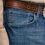Close-up of a person wearing Mens Wrangler 20X No. 42 Vintage Bootcut Jean in premium denim blue, paired with a patterned brown leather belt featuring a decorative metal buckle and a portion of a light-colored shirt visible on the left.