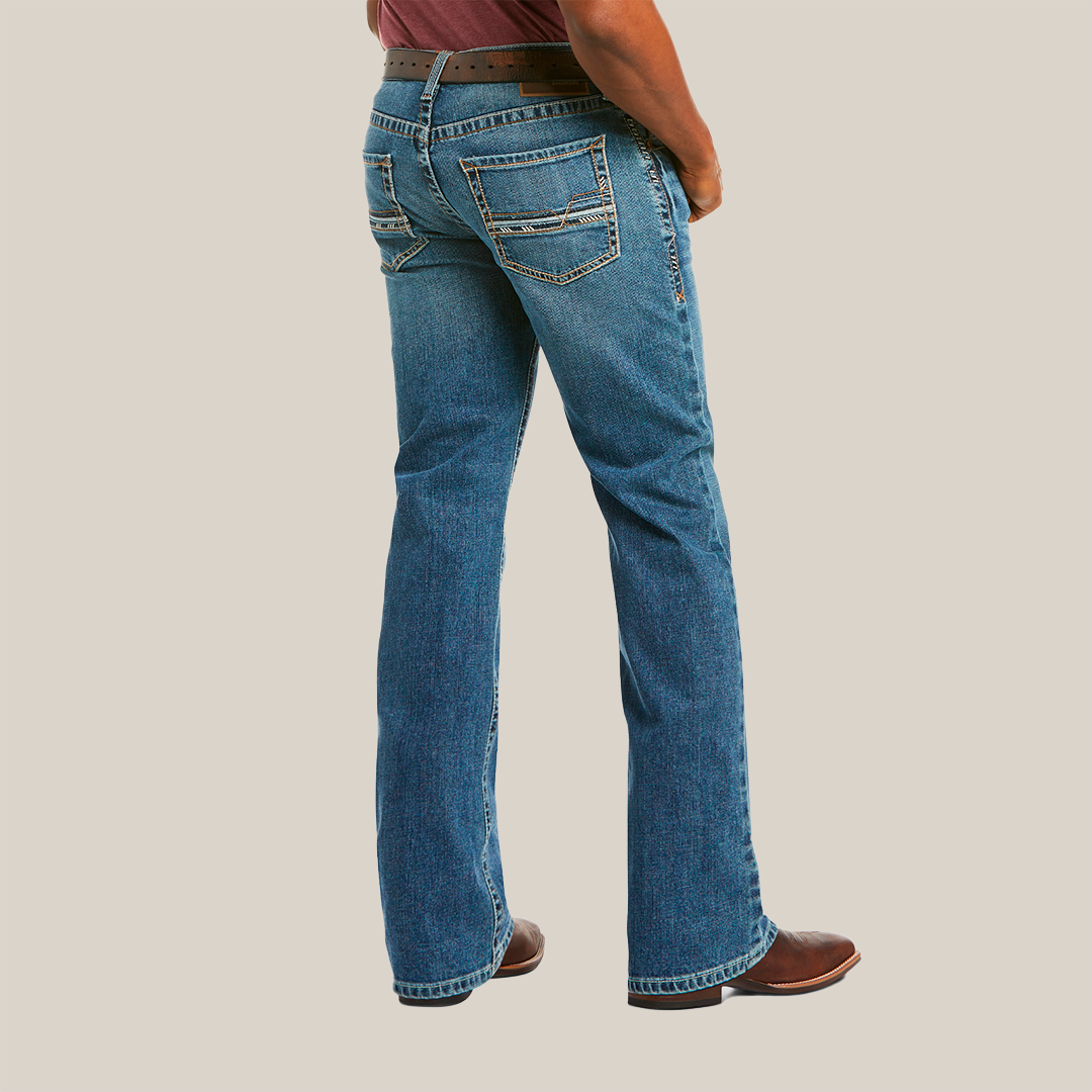 A person is wearing M5 Slim Stretch Stillwell Stackable Straight Leg Jean - Fargo, featuring premium cotton in blue with brown stitching, paired with a dark belt and brown shoes. The image shows the back view from the waist down against a plain white background, highlighting two decorative-stitched back pockets.