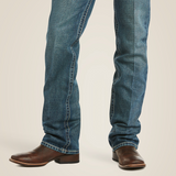 A person is wearing a dark gray t-shirt, Ariat M5 Slim Straight Leg Jeans in blue denim (Gulch - 10014010), and brown leather boots. They have a brown belt with a silver buckle and are standing against a white background with one hand in their pocket.
