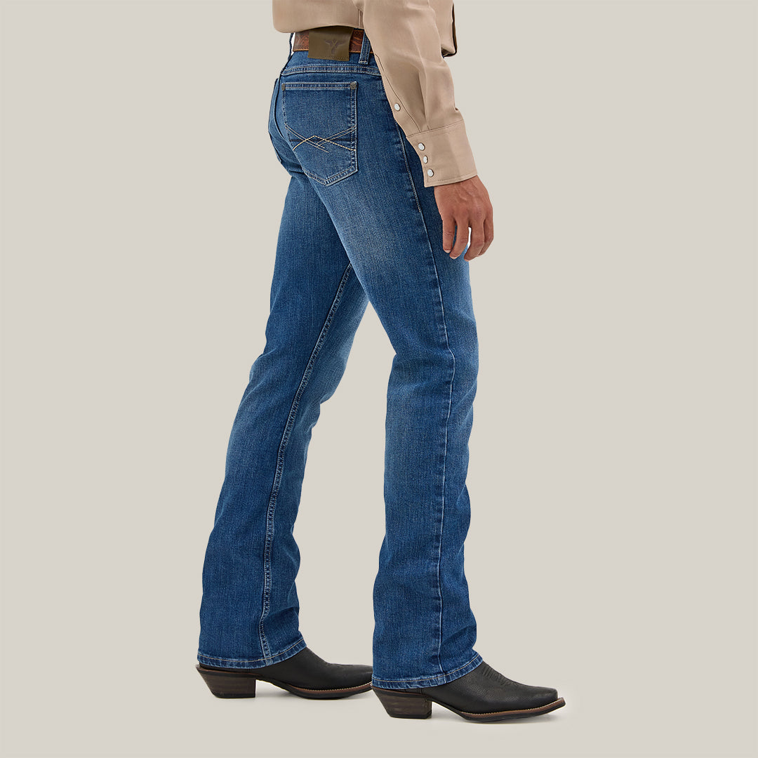 A person in blue denim jeans and black boots is shown from the side against a plain background, wearing a beige shirt with white buttons. The jeans, Mens Wrangler 20X No. 42 Vintage Bootcut Jean - 112338537, feature stitching details and a distinctive Wrangler patch on the back pockets.