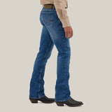 A person in blue denim jeans and black boots is shown from the side against a plain background, wearing a beige shirt with white buttons. The jeans, Mens Wrangler 20X No. 42 Vintage Bootcut Jean - 112338537, feature stitching details and a distinctive Wrangler patch on the back pockets.