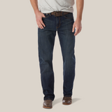 A person wearing a gray t-shirt, Wrangler 20X No. 33 Extreme Appleby Dark Wash Relaxed Straight Leg Jeans - 1033MWXAB, and brown leather shoes stands on a white background. Their right hand is in a pocket, highlighting the premium denim style with a coordinating brown belt.