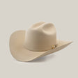The 500X Superior Belly, a beige cowboy hat made from 100% belly beaver fur, features a wide brim and a decorative band with a metallic embellishment. Displayed on a smooth light gray surface, it is slightly tilted to highlight its premium craftsmanship.