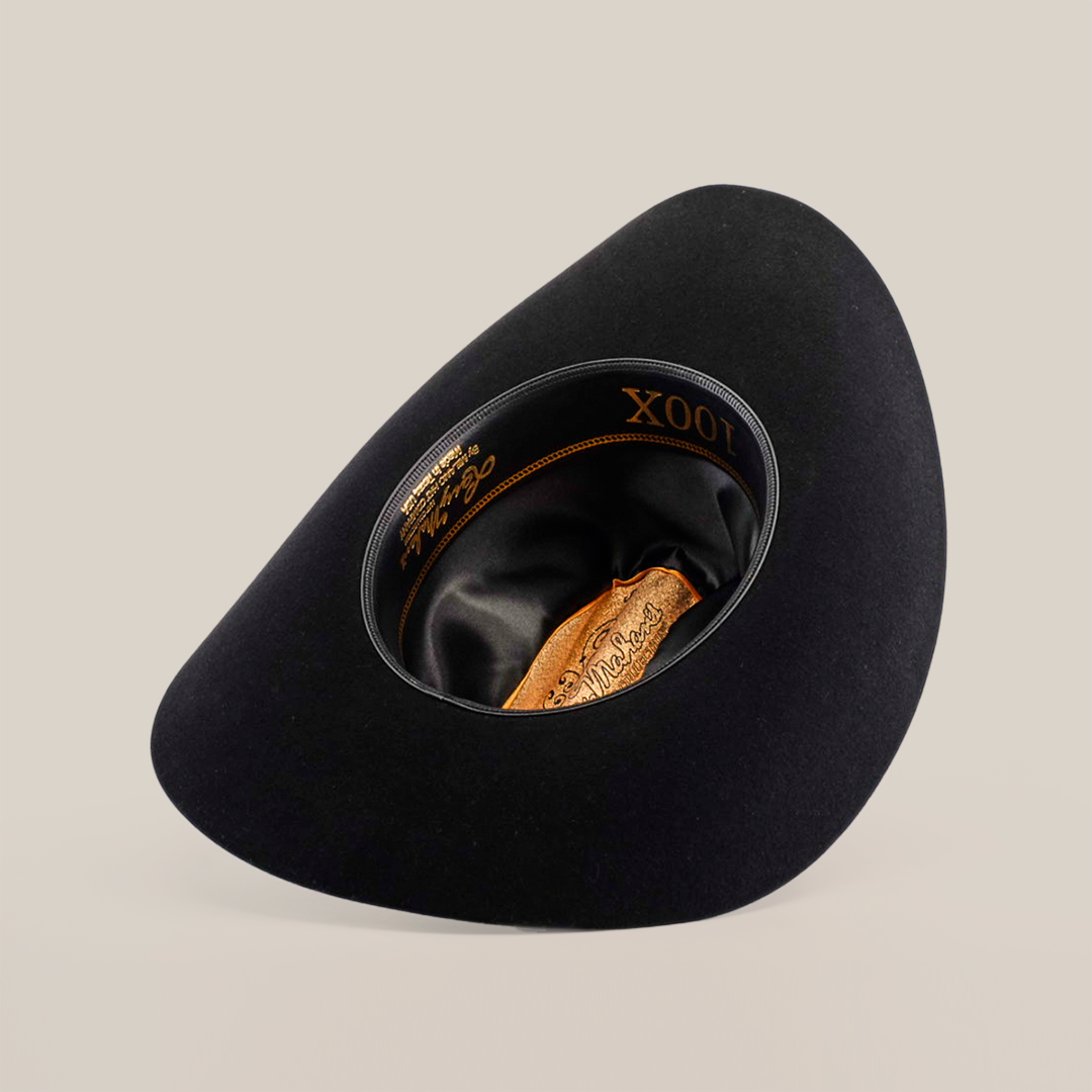 The 100X Independecia Black is a black, wide-brimmed hat turned upside down, showcasing its satin lining and leather headband. This epitome of western headwear features elegant gold detailing inside, including the distinguished Larry Mahan 100X.