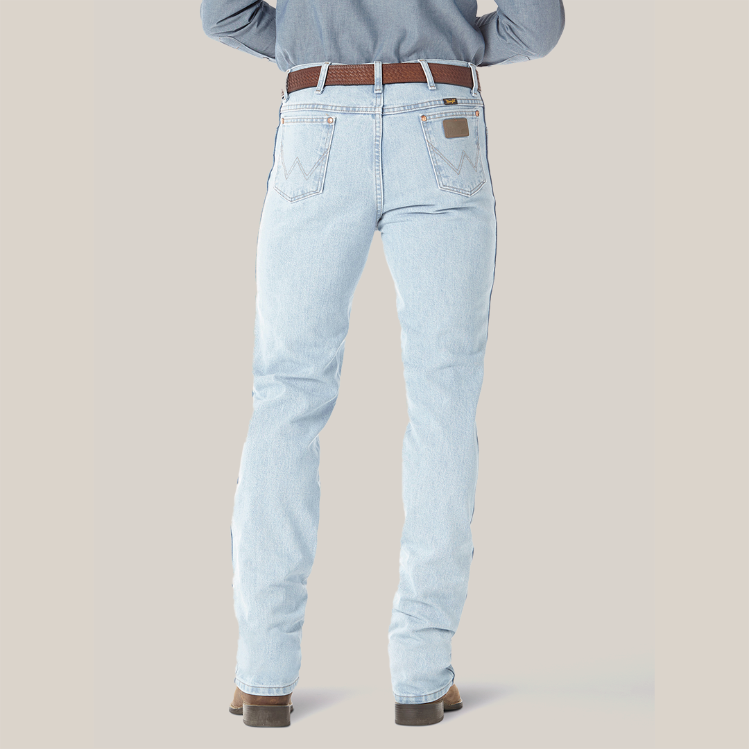A person is seen from the rear in Wrangler Cowboy Cut Slim Fit Jean - 0936GBH, paired with a brown leather belt and tan boots. These premium jeans showcase distinctive stitching on the back pockets. The stance is slightly apart against a light background.