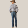 A modern cowboy stands with his back to the camera, wearing a plaid shirt, Ariat M7 Slim Ezra Straight Jeans - Brighton (10045388), brown boots, and a cowboy hat.