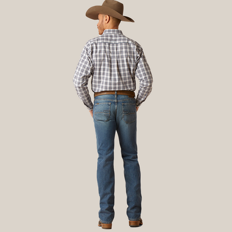 A modern cowboy stands with his back to the camera, wearing a plaid shirt, Ariat M7 Slim Ezra Straight Jeans - Brighton (10045388), brown boots, and a cowboy hat.