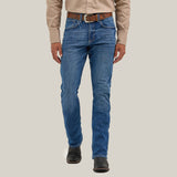 A person in a beige long-sleeve shirt, blue jeans, and a brown belt stands before a plain background. They wear Mens Wrangler 20X No. 42 Vintage Bootcut Jeans with the iconic patch, hands at their sides, while black boots complete the outfit.