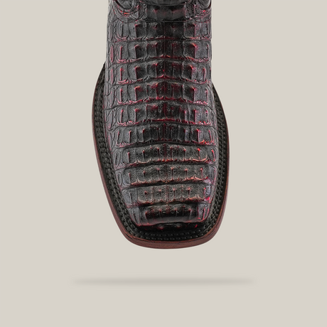 A close-up of the Exotic Caiman Hornback - Black Cherry boot showcases its black and red crocodile pattern with a square toe, crafted from genuine caiman leather. White stitching around the sole highlights expert craftsmanship against a pristine white background.
