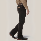 A person sporting a brown button-up shirt and dark jeans with a visible brown belt exudes rugged style. The Wrangler Cowboy Cut Slim Fit Jean - 0936KCL pairs perfectly with brown cowboy boots as they stand with one foot forward, emphasizing the side profile.