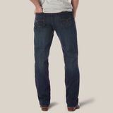 A person poses against a white backdrop wearing the Wrangler 20X No. 33 Extreme Appleby Dark Wash Relaxed Straight Leg Jeans, featuring orange stitching and styled with brown shoes, embodying a laid-back essence with hands in back pockets and a light gray shirt.