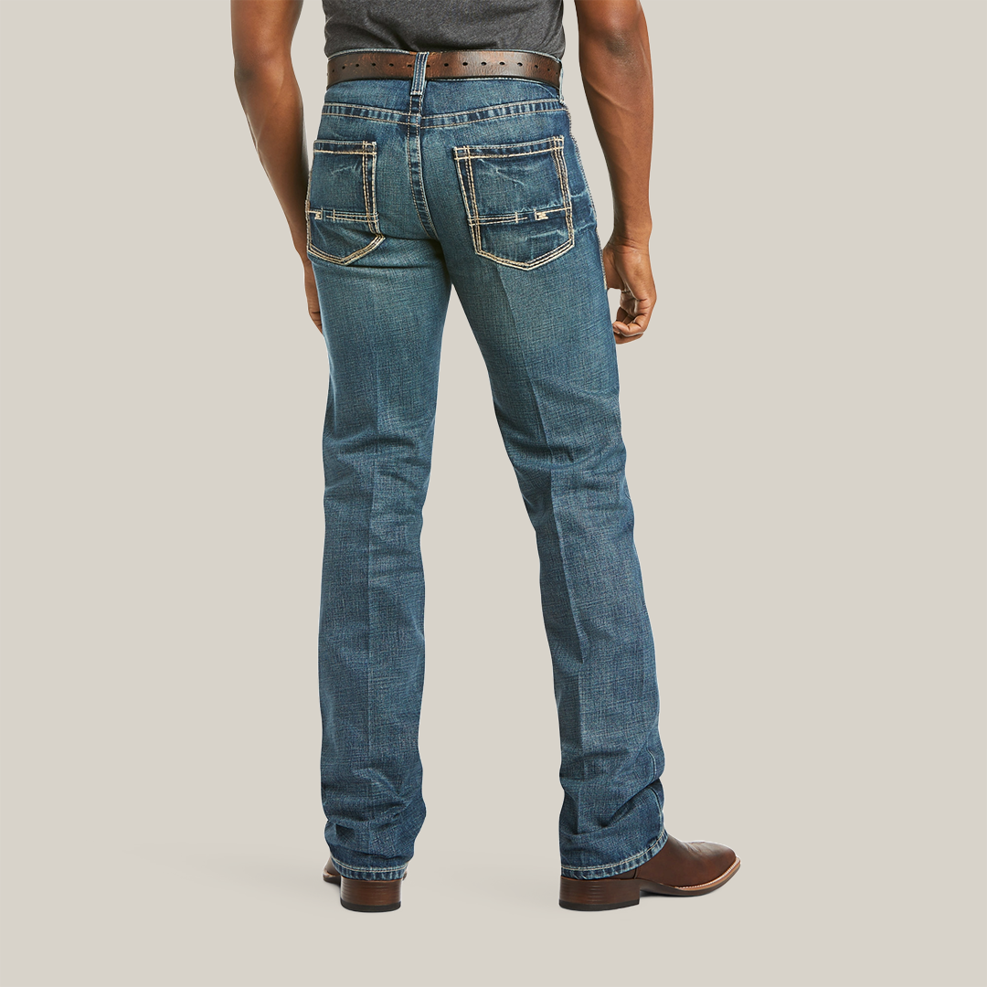 A person stands back to the camera in M5 Slim Boundary Stackable Straight Leg Jeans - Gulch - 10014010 by Ariat, featuring premium cotton blue denim with white stitching. Paired with a brown belt and shoes, two back pockets, boot-cut hems, and a tucked dark shirt for a polished look.
