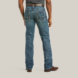 A person stands back to the camera in M5 Slim Boundary Stackable Straight Leg Jeans - Gulch - 10014010 by Ariat, featuring premium cotton blue denim with white stitching. Paired with a brown belt and shoes, two back pockets, boot-cut hems, and a tucked dark shirt for a polished look.
