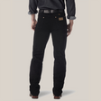 A person faces away from the camera, wearing a dark long-sleeve shirt with rolled-up sleeves, black Wrangler Cowboy Cut Slim Fit Jeans - 0936WBK made from premium denim, and brown leather shoes. A belt is partially visible above the waistband of their jeans.