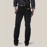 A person faces away from the camera, wearing a dark long-sleeve shirt with rolled-up sleeves, black Wrangler Cowboy Cut Slim Fit Jeans - 0936WBK made from premium denim, and brown leather shoes. A belt is partially visible above the waistband of their jeans.