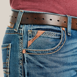 A close-up of someone wearing M5 Slim Stretch Stillwell Stackable Straight Leg Jeans - Fargo shows premium cotton blue denim with detailed stitching and a pocket logo. A brown leather belt with metal holes loops through the belt loops, effortlessly complementing the maroon shirt.