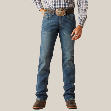 Dressed in the M7 Slim Ezra Straight Jean - Brighton, a person stands with hands at sides. The jeans have a slightly faded look, ideal for a modern cowboy, paired with brown cowboy boots and a large decorative belt buckle.