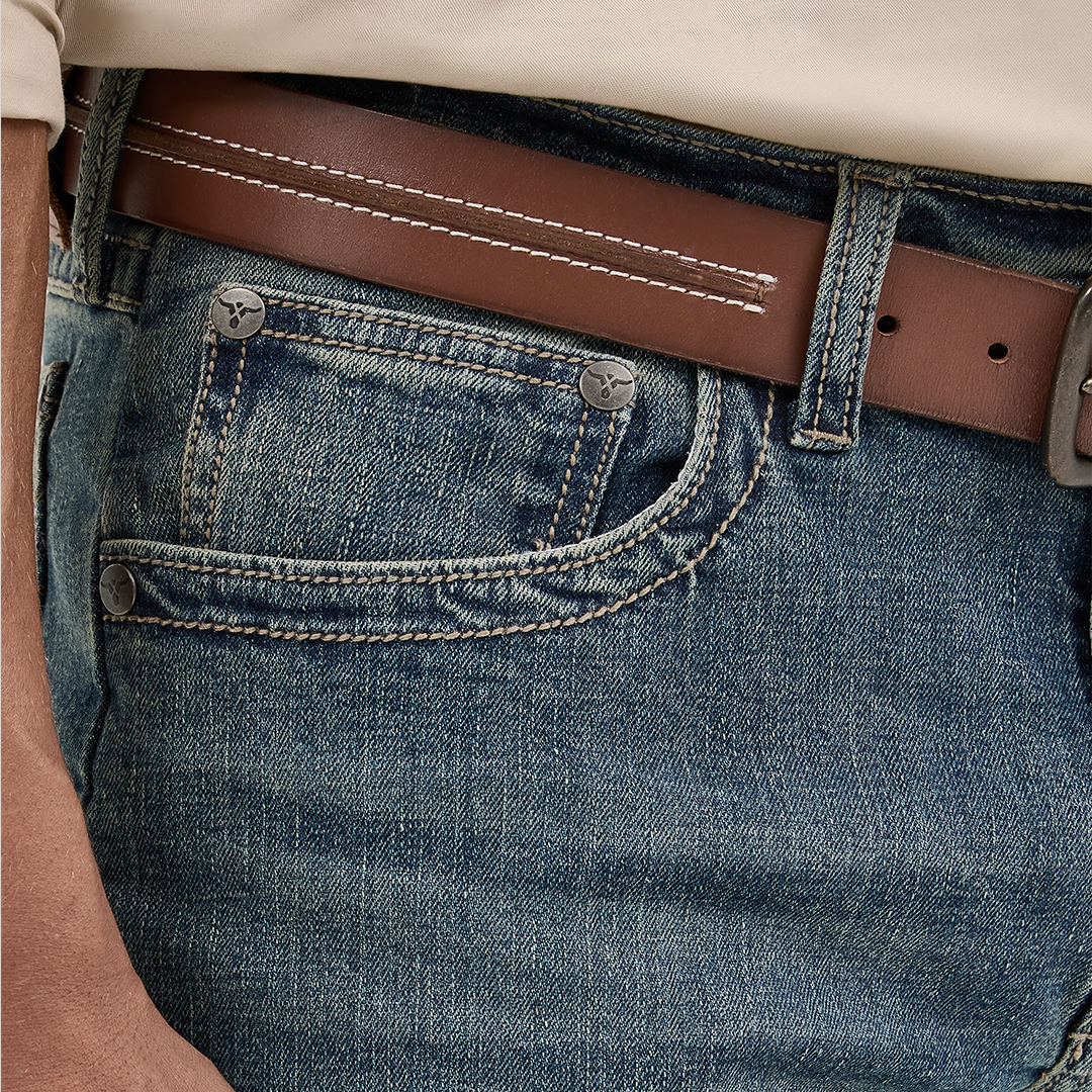A hand casually rests in the pocket of Wrangler Mens 20X Extreme Relaxed Fit Jeans - 1044MWXMA, featuring detailed stitching. Theyre paired with a brown leather belt with white stitching, while a beige shirts hem subtly peeks out.