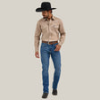 A man in a cowboy hat stands against a plain white background, wearing a beige long-sleeved shirt and Mens Wrangler 20X No. 42 Vintage Bootcut Jeans crafted from premium denim. Dark boots complete his rugged look as he gazes downward, embodying timeless Western style.